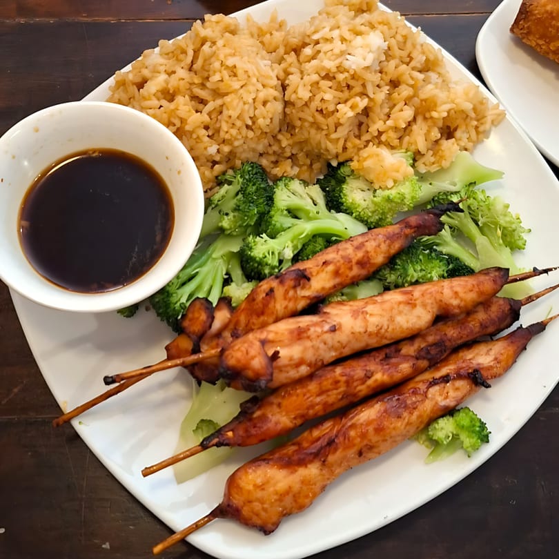 Chicken teriyaki skewers with fried rice, broccoli, and a side of soy sauce on a white plate.