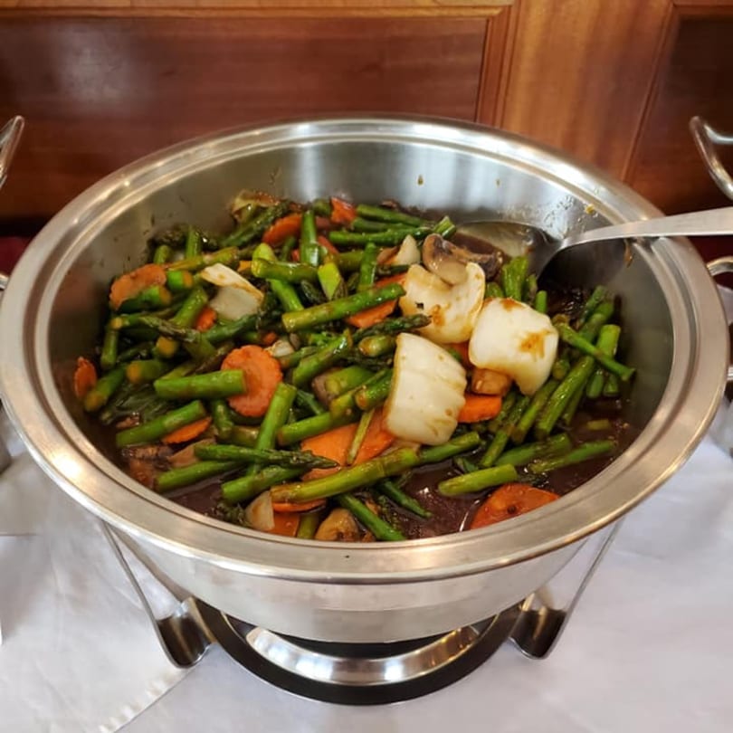 A silver chafing dish filled with stir-fried asparagus, carrots, onions, and mushrooms in a dark sauce.