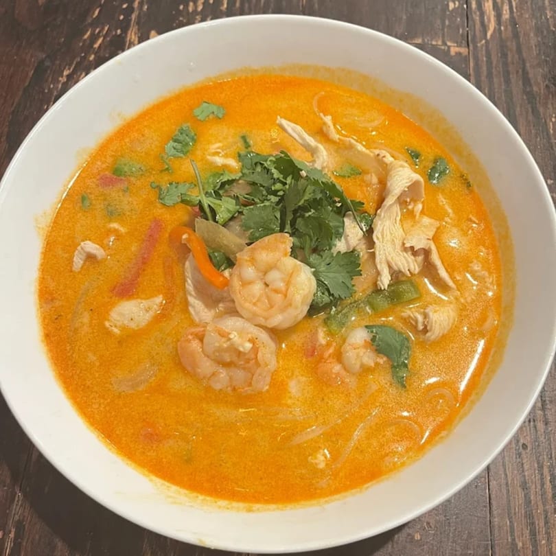 A vibrant orange soup with shrimp, chicken, and cilantro in a white bowl on a wooden table.